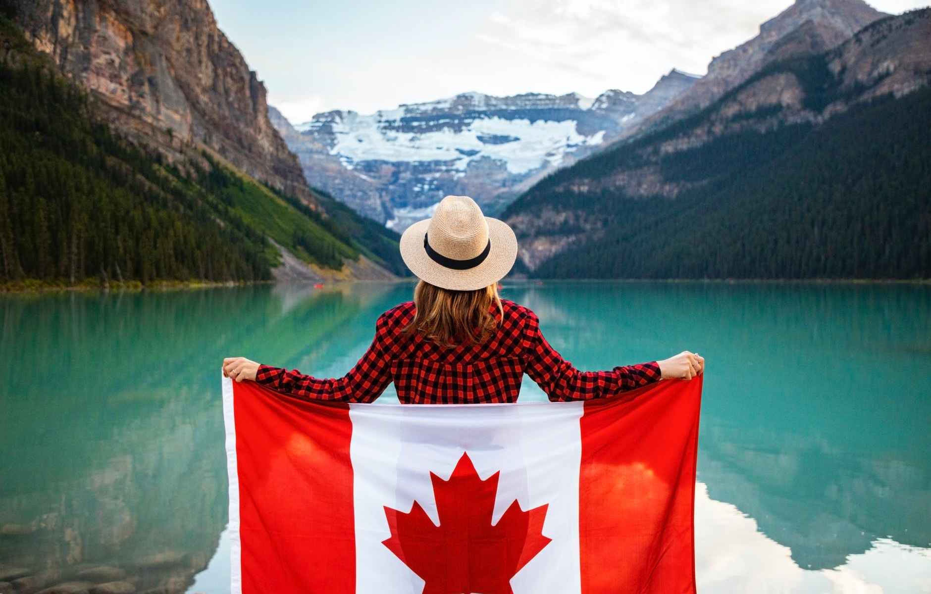 woman wearing red and black checkered dress shirt and beige fedora hat holding canada flag looking at lake